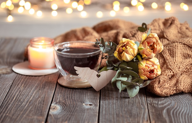 Festliches Stillleben mit einem Getränk in einer Tasse, Blumen und einem Strickartikel auf einer Holzoberfläche vor einem unscharfen Hintergrund mit Bokeh.