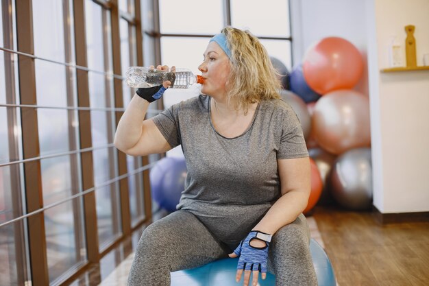 Fette Frau, die nährt, Fitness. Dame, die auf einem Fitball sitzt und ein Wasser trinkt.