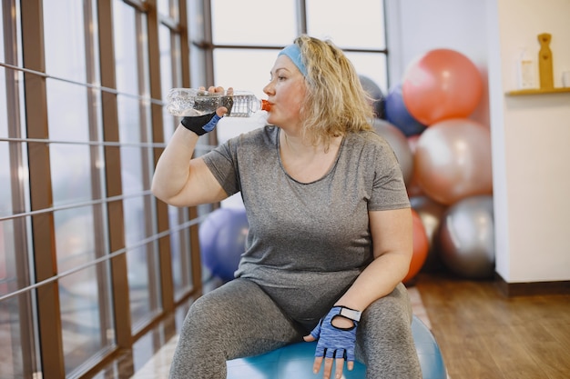 Fette Frau, die nährt, Fitness. Dame, die auf einem Fitball sitzt und ein Wasser trinkt.