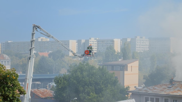 Feuerwehrleute auf Plattformwagen gehen auf Dach des Hauses in Brand. Blick auf Feuerwehrleute, die versuchen, Feuer aus brennendem Gebäude in Flammen und Dämpfen zu löschen. Männer stoppen Smog und Rauch aus dem Haus.