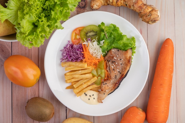 Fischsteak mit Pommes Frites, Kiwi, Salat, Karotten, Tomaten und Kohl in einer weißen Schale.