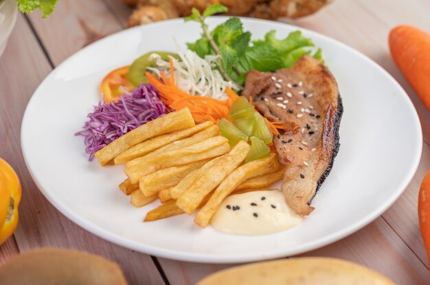 Fischsteak mit Pommes Frites, Kiwi, Salat, Karotten, Tomaten und Kohl in einer weißen Schale.