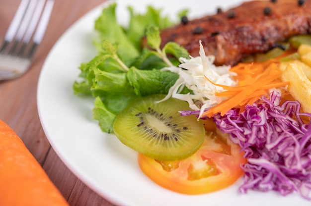 Fischsteak mit Pommes Frites, Kiwi, Salat, Karotten, Tomaten und Kohl in einer weißen Schale.