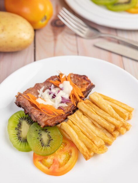 Fischsteak mit Pommes Frites, Kiwi, Salat, Karotten, Tomaten und Kohl in einer weißen Schale.