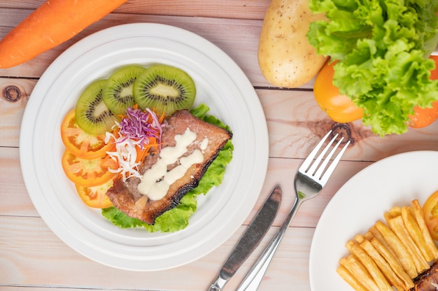 Fischsteak mit Pommes Frites, Kiwi, Salat, Karotten, Tomaten und Kohl in einer weißen Schale.