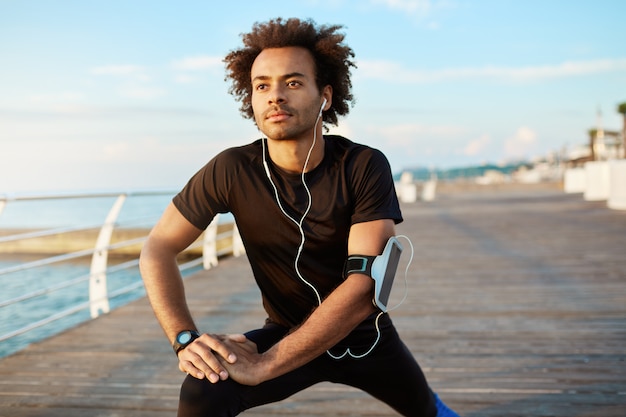 Fit afroamerikanischer Jogger mit buschiger Frisur, die seine Muskeln vor dem Laufen aufwärmt. Mannathlet in der schwarzen Sportbekleidung, die Beine mit Stretchübung auf hölzernem Pier mit weißen Kopfhörern auf streckt.