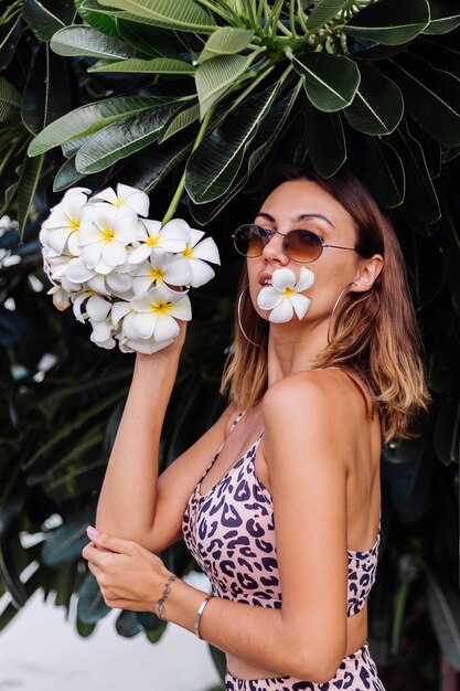 Fit europäische stilvolle Frau in Sonnenbrille, Leopardenoberteil und Shorts auf Blumenbaum in Thailand tragend