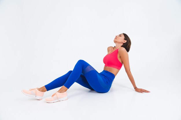 Fit gebräunte sportliche Frau mit Bauchmuskeln, Fitnesskurven, trägt Top und blaue Leggings auf weiß