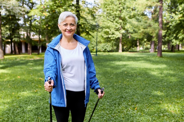 Fit glückliche grauhaarige reife Frau in Sportbekleidung genießen gesundheitsfördernde körperliche Aktivität unter Verwendung von Wanderstöcken, die freudigen Gesichtsausdruck erregen, frische Luft in wilder Natur atmen, lächeln