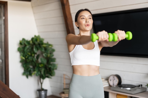 Fitness asiatisches Mädchen, das zu Hause im Wohnzimmer trainiert, Hanteln heben und Activewear für Sport tragen ...