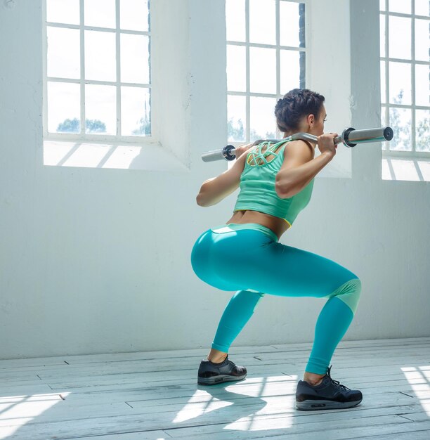 Fitness-Frau von hinten in azurblauer Sportbekleidung, die Kniebeugen mit Langhantel auf ihren Schultern macht.