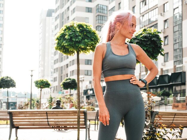 Fitness lächelnde Frau in grauer Sportkleidung mit rosa Haaren Junges schönes Modell mit perfektem KörperFrau posiert auf der Straße HintergrundFröhlich und glücklich Stretching vor dem Training