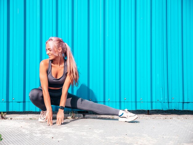 Fitness lächelnde Frau in grauer Sportkleidung mit rosa Haaren Junges schönes Modell mit perfektem KörperFrau posiert auf der Straße in der Nähe der blauen Wand Fröhlich und glücklich Stretching vor dem Training