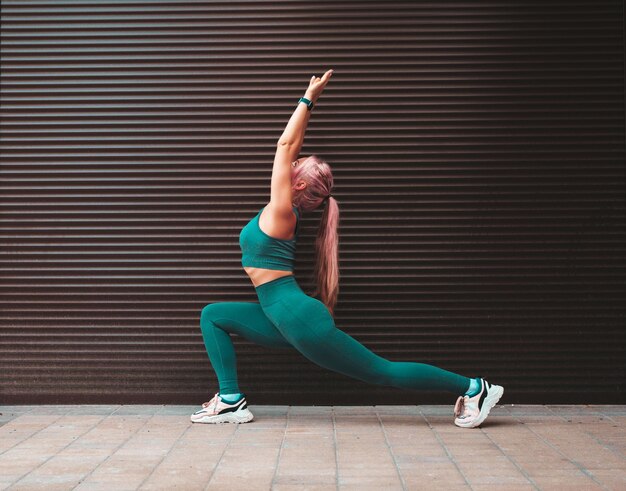 Fitness lächelnde Frau in grüner Sportkleidung mit rosa Haaren Junges schönes Modell mit perfektem Körper Weibliche Pose auf der Straße in der Nähe der Rollladenwand Dehnung vor dem Training