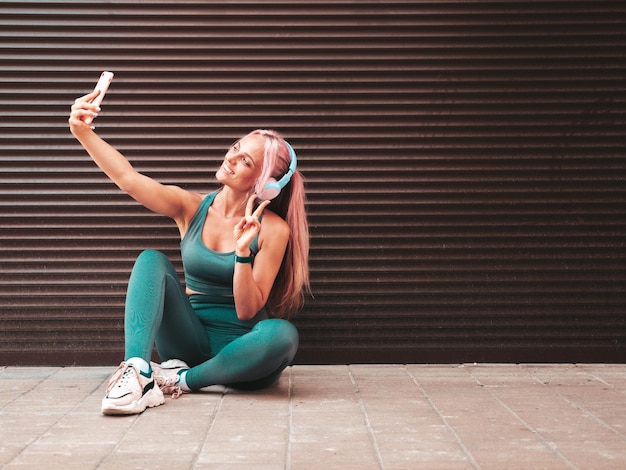 Fitness lächelnde Frau in grüner Sportkleidung mit rosa HaarenJunges schönes Modell mit perfektem KörperFrau auf der Straße in der Nähe von RollladenwandMusik hören in drahtlosen KopfhörernSelfie machen