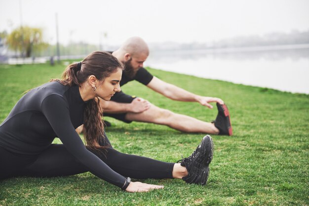 Fitness-Paar, das draußen im Park nahe dem Wasser streckt. Junger bärtiger Mann und Frau, die zusammen am Morgen trainieren