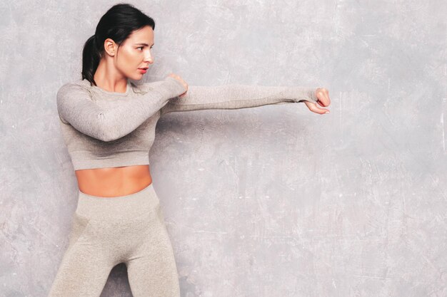 Fitness selbstbewusste Frau in Sportkleidung Sexy junges schönes Modell steht in einem Gestell. Frau posiert in der Nähe einer grauen Wand im Studio. Sie übt Kraftschläge beim Boxen während des Trainings