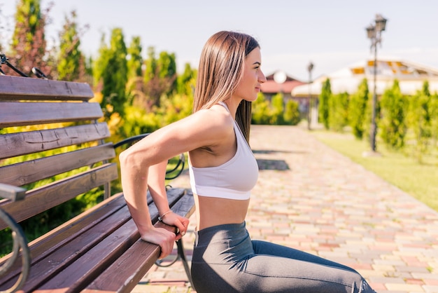 Fitness-, Sport-, Trainings-, Park- und Lifestyle-Konzept. Junge lächelnde Frau, die Liegestütze auf Bank im Freien tut