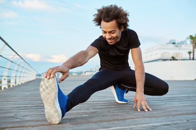 Fitness und Motivation. Freudiger und lächelnder dunkelhäutiger Athlet, der sich morgens auf dem Pier ausdehnt. Sportlicher afroamerikanischer Mann mit buschigem Haar, das seine Beine aufwärmt