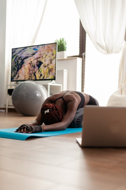 Fitte Frau in Sportkleidung, die sich auf der Yogamatte entspannt und Online-Trainingsübungen mit dem Laptop im Wohnzimmer für das Wohlbefinden ansieht
