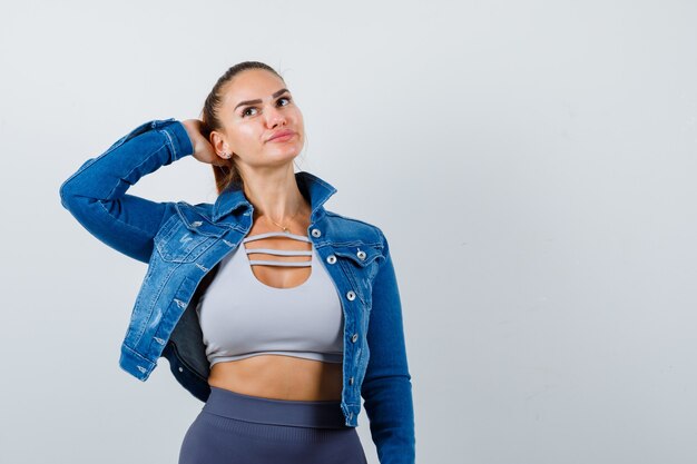 Fitte Frau mit der Hand auf dem Kopf, die an etwas in bauchfreiem Top, Jeansjacke, Leggings und nachdenklichem Aussehen denkt. Vorderansicht.