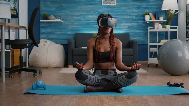 Fitte schwarze Frau mit Virtual-Reality-Headset, während sie auf Yoga-Karte sitzt