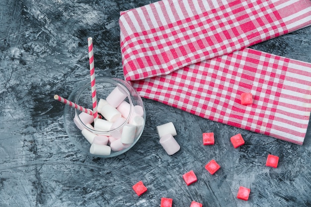 Flach gelegte Marshmallows und Zuckerrohre im Glas mit Süßigkeiten und roter Gingham-Tischdecke auf dunkelblauer Marmoroberfläche. horizontal