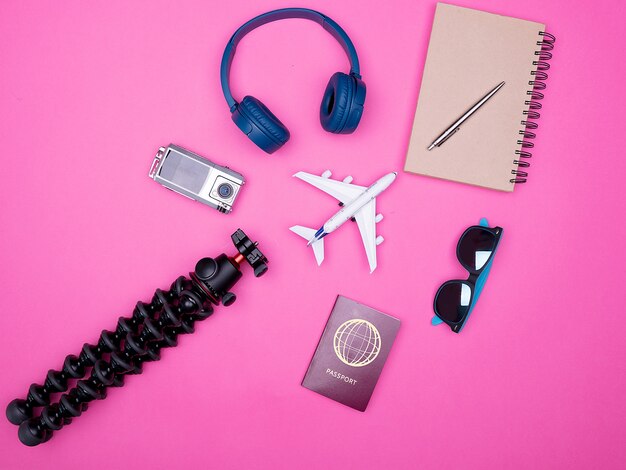 Flache Draufsicht auf das Zubehör von Reisendenfotografen auf rosafarbenem Hintergrund. Notizbuch, Reisepass, Stativ, Kopfhörer und Sonnenbrille