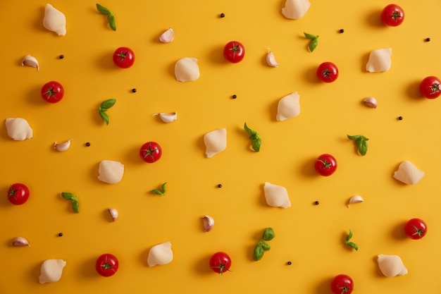 Flache Draufsicht auf Nudelschalen aus Hartweizen, roten Kirschtomaten, Basilikum und Knoblauch zur Zubereitung eines italienischen Gerichts. Gemüse und Gewürze auf gelbem Hintergrund. Zutaten kochen. Detox Essen