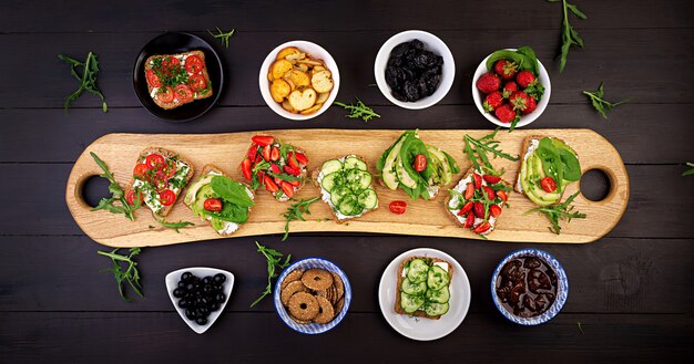 Flache Lage der gesunden vegetarischen Tischdekoration. Sandwiches mit Tomaten, Gurken, Avocado, Erdbeere, Kräutern und Oliven, Snacks. Sauberes Essen, veganes Essen
