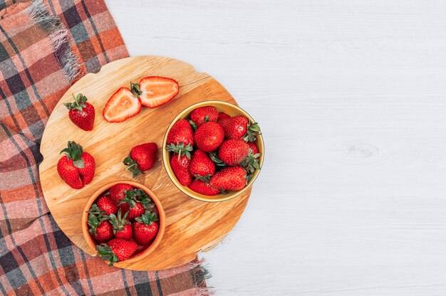 Flache legen Bündel Erdbeeren in gelben Schalen mit geteilter halber Erdbeere auf weißem Holz und strukturiertem Stoffhintergrund. horizontal