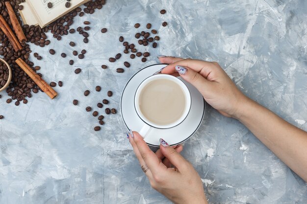 Flache weibliche Hände, die eine Tasse Kaffee mit Kaffeebohnen, Zimtstangen, Buch auf grauem Gipshintergrund halten. horizontal