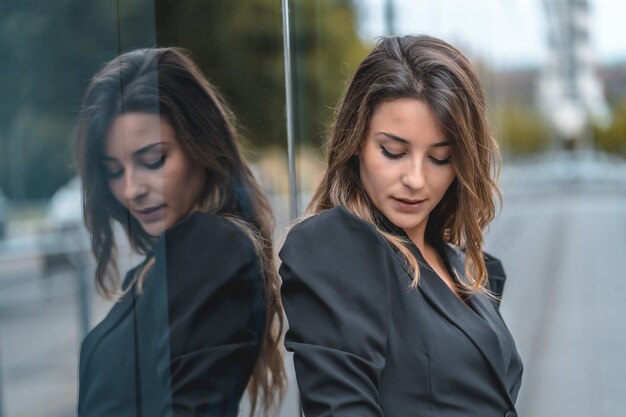 Flacher Fokus einer jungen Frau in einem schwarzen Kleid, die in der Nähe eines modernen Gebäudes mit Glaswänden posiert