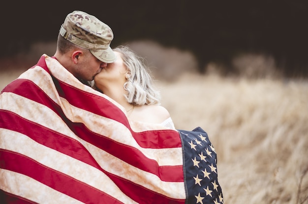 Flacher Fokusschuss eines amerikanischen Soldaten, der seine liebende Frau küsst, während er in eine amerikanische Flagge gehüllt wird