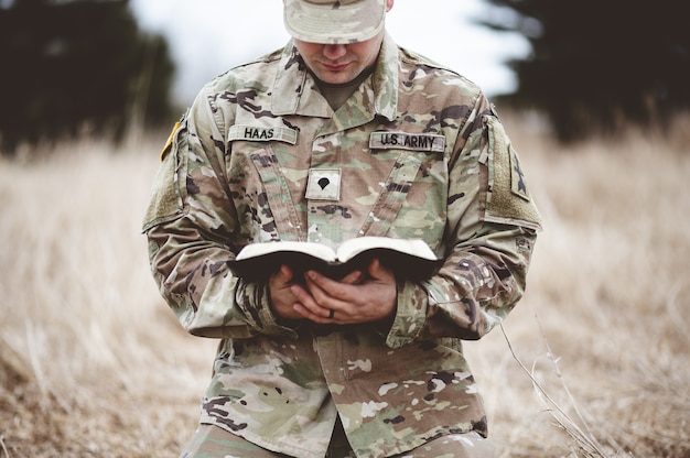 Flacher Fokusschuss eines jungen Soldaten, der auf einem trockenen Gras kniet, während er die Bibel liest