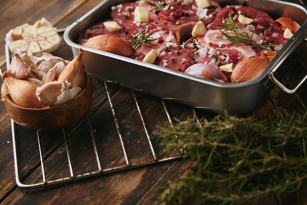 Fleisch in einer Stahlpfanne mit Gewürzen: Knoblauch, Rosmarin, Zwiebeln; bereit, auf dickem Grunge gebürstetem Holztisch zu kochen