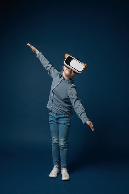 Fliegen wie ein Flugzeug. Kleines Mädchen oder Kind in Jeans und Hemd mit Virtual-Reality-Headset-Brille lokalisiert auf blauem Studiohintergrund. Konzept der Spitzentechnologie, Videospiele, Innovation.
