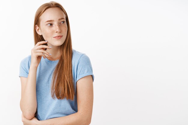 flirty faszinierte und interessierte gut aussehende erwachsene rothaarige Frau mit Sommersprossen im blauen T-Shirt, die sich mit leichtem sinnlichem Lächeln nach rechts dreht und mit Verlangen und Zuneigung blinzelt