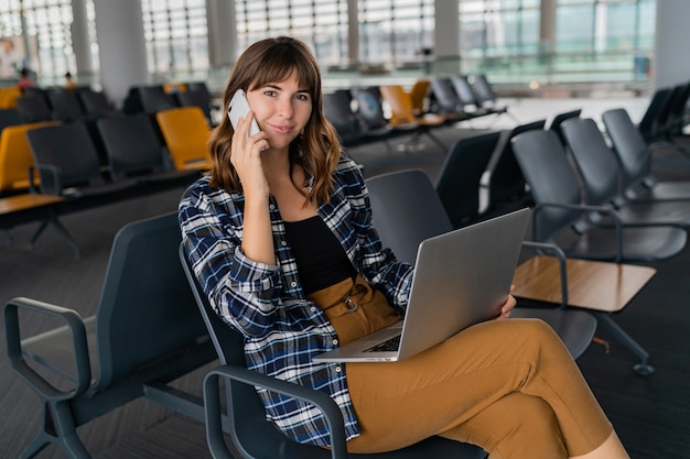 Flughafen junge Passagierin mit Smartphone und Laptop sitzen in der Terminalhalle, während sie auf ihren Flug wartet