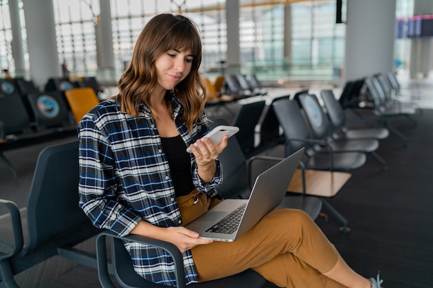 Flughafen junge Passagierin mit Smartphone und Laptop sitzen in der Terminalhalle, während sie auf ihren Flug wartet