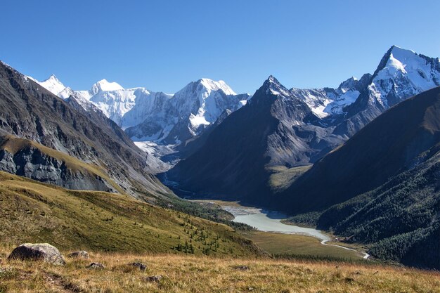 Fluss, umgeben von felsigen Hügeln, die unter dem Sonnenlicht und einem blauen Himmel mit Schnee bedeckt sind