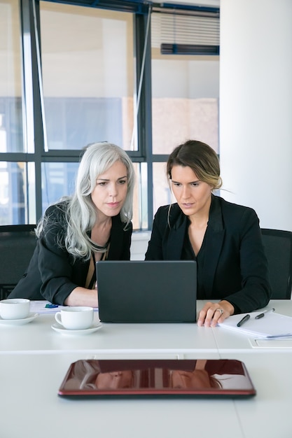 Fokussierte aufgeregte Geschäftsfrauen, die Laptopanzeige beim Sitzen am Tisch mit Tassen Kaffee im Büro betrachten. Vorderansicht. Teamwork und Kommunikationskonzept