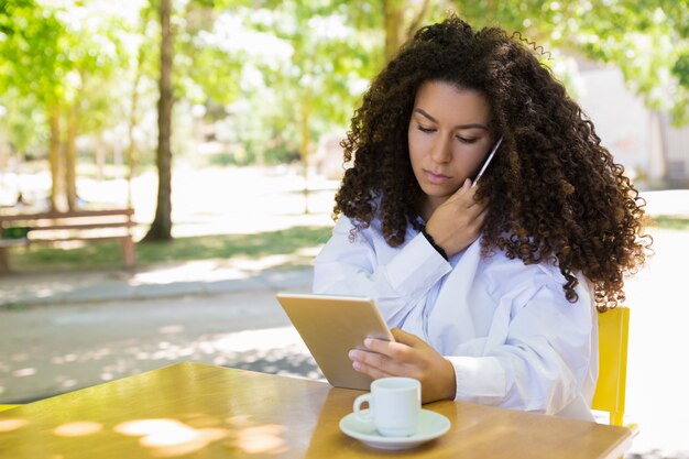 Fokussierte Dame, die um Telefon ersucht und Tablette Café im im Freien verwendet