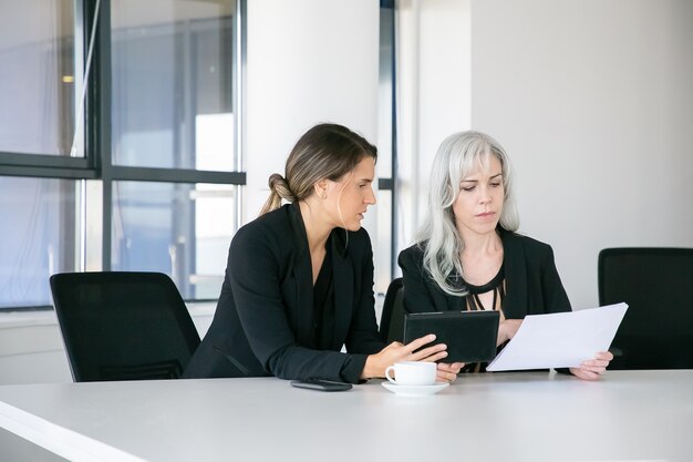 Fokussierte Fachleute, die gemeinsam Berichte analysieren. Zwei Geschäftsfrauen sitzen zusammen, lesen Dokumente, benutzen Tablette und sprechen. Teamwork-Konzept