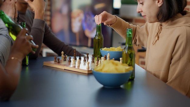 Fokussierte Frau, die mit Freunden strategische Brettspiele spielt, Spaß mit Schachmatch und Wettbewerb zu Hause hat. Fröhliche Menschen genießen gemeinsam das Gameplay und servieren Bier. Handschuss. Nahansicht.
