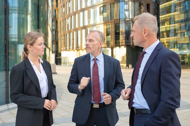 Kostenloses Foto fokussierte geschäftskollegen, die büroanzüge tragen, sich im freien treffen, stehen und mit stadtgebäuden im hintergrund sprechen. konzept der unternehmenskommunikation