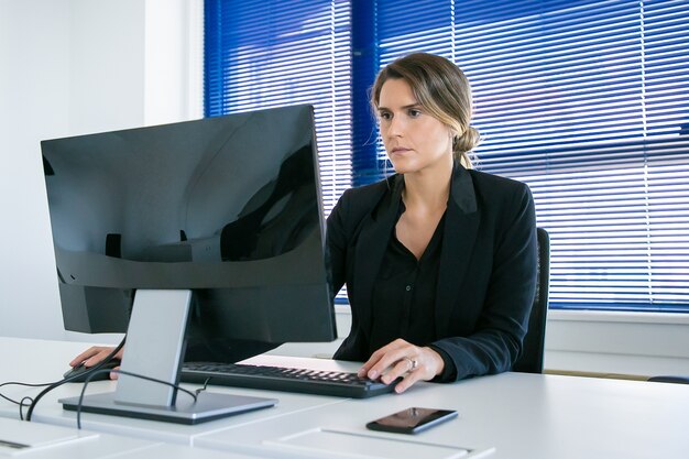 Fokussierte junge Geschäftsdame, die in ihrem Büro arbeitet, Computer am Arbeitsplatz benutzt und Anzeige betrachtet. Mittlerer Schuss. Konzept für digitale Kommunikation oder Unternehmensleiter