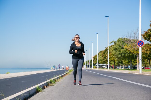 Fokussierte reife Frau in der Fitnesskleidung, die entlang des Flussufers draußen joggt und Morgenlauf genießt. Vorderansicht in voller Länge. Aktives Lifestyle-Konzept