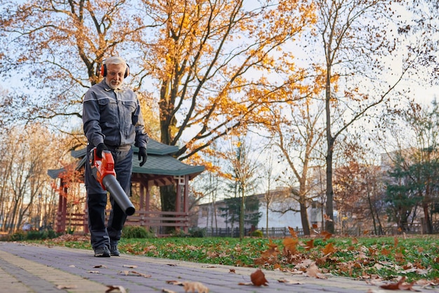 Kostenloses Foto fokussierter älterer mann, der im oktober den bürgersteig mit einem laubbläser reinigt und einen lächelnden bärtigen aus der tiefe betrachtet