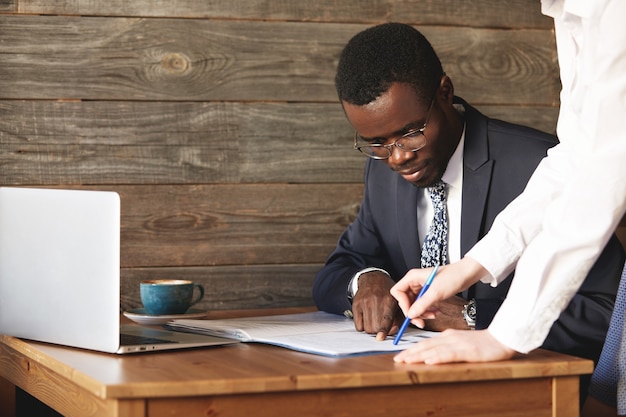 Fokussierter afroamerikanischer Geschäftsmann, der Papiere mit seinem persönlichen Assistenten im weißen Hemd prüft